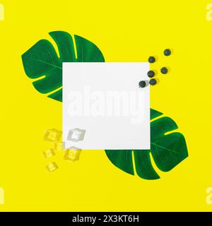 Mise en page créative faite avec des feuilles tropicales vertes, des glaçons, des bleuets et un espace de copie de note de carte de papier sur fond jaune. Concept estival minimal. Banque D'Images