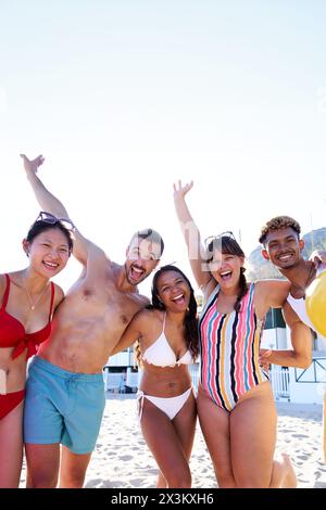 Portrait de cinq amis sur la plage s'amusant à regarder la caméra souriant et levant les bras. Banque D'Images