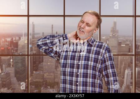 Bel homme caucasien mature a un mal de cou. Arrière-plan de fenêtres à carreaux avec vue paysage urbain. Banque D'Images