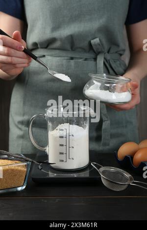 Femme ajoutant de la levure chimique dans la tasse à mesurer à la table en bois noir, gros plan Banque D'Images