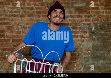 Jeune homme latino (28 ans) à vélo avec un panier sourit alors qu'il se rend au travail. Espace de copie, fond de brique. Travaux conceptuels Banque D'Images