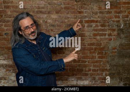 Homme latino-américain (44) avec de longs cheveux gris, veste de jeans bleus pointe avec les deux bras sur son côté gauche. Copier l'espace. Fond de brique. Concept de style de vie Banque D'Images