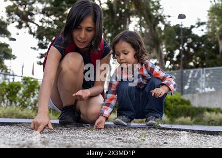 Maman apprend à son fils latino-américain à chercher de petites pierres dans le sable dans une aire de jeux pour enfants. Concept et enseignement de la fête des mères Banque D'Images