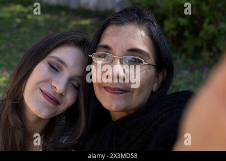 Mère latino-américaine (50) et fille (24) prennent un selfie dans le parc, la fille s'appuie amoureusement sur l'épaule de sa mère fermant les yeux. Mère » Banque D'Images