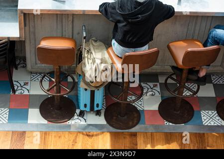 Vue arrière d'une femme assise dans un tabouret de bar, appuyée contre le comptoir en bois, avec son chariot attend le départ de leur vol de CDG airpor Banque D'Images