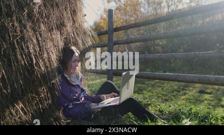 Une femme aux cheveux longs est assise paisiblement dans l'herbe verte luxuriante, tapant sur un ordinateur portable sous une pile de foin. Freelance travaille à distance dans la campagne d'automne au coucher du soleil. Voyage, liberté, mode de vie actif Banque D'Images