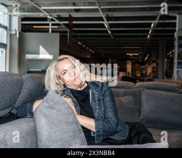 Joyeuse dame aux cheveux argentés dans une veste en denim foncé nichée sur un canapé charbon de bois Banque D'Images