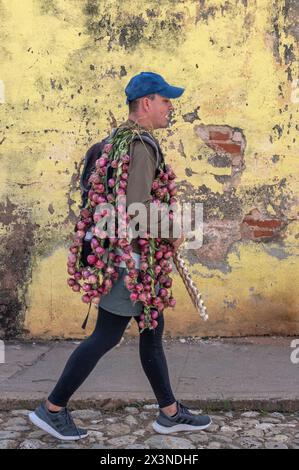 Un vendeur de rue marchant devant un mur peint en jaune, altéré, transportant des oignons et de l'ail dans les ruelles de la vieille ville, Trinidad, Cuba. Banque D'Images