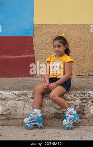 Une jeune fille locale s’est assise sur le bord du trottoir surélevé devant un mur coloré, portant ses patins à roulettes dans la vieille ville de Trinidad, Cuba Banque D'Images