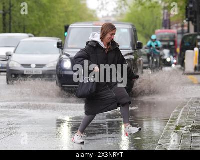 Les piétons sautent au-dessus d'un bassin d'eau de pluie qui s'est accumulé sur la section ouest de la route Euston, adjacente à la jonction avec Woburn place, Londres. Date de la photo : dimanche 28 avril 2024. Banque D'Images