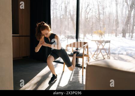 Femme mince sportive faisant une fente sur une jambe à l'aide d'une chaise dans le salon. Vue latérale d'une femme de fitness motivée effectuant une pose de fente sur une jambe sur une chaise Banque D'Images