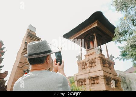 Un homme adulte avec un chapeau gris capture une photo du temple hindou balinais traditionnel avec un arrière-plan bokeh ou flou Banque D'Images