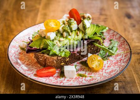 Gros plan d'avocat avec fromage feta, tomates, pignons de pin, pesto et salade sur pain brun sur une assiette sur une table en bois Banque D'Images