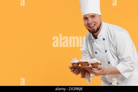 Heureux confiseur professionnel en uniforme tenant de délicieux cupcakes sur fond jaune. Espace pour le texte Banque D'Images