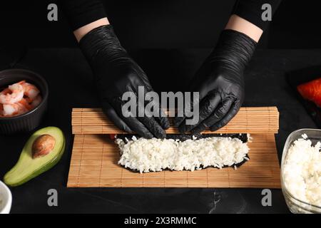 Chef en gants enveloppant le rouleau de sushi à la table texturée sombre, gros plan Banque D'Images