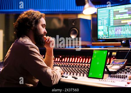 Ingénieur du son qualifié regarde l'affichage de la maquette sur la tablette avant de mixer et de mastering des pistes sur la console stéréo électronique. Le technicien utilise un logiciel audio numérique pour enregistrer et éditer ses chansons. Banque D'Images