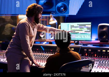 Musicien qualifié travaillant avec un ingénieur du son pour éditer de nouvelles pistes enregistrées dans la salle de contrôle, en appuyant sur les boutons et les faders sur le bureau du tableau de bord. Équipe d'artiste et producteur créant de la musique en studio. Banque D'Images