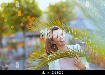 Portrait de femme élégante heureuse en robe rose et veste blanche dans la ville près de l'arbre tropical. Banque D'Images