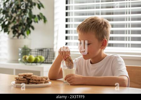 Garçon souriant alors qu'il trempe un biscuit fait maison dans un verre de lait, profitant de sa collation Banque D'Images