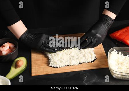 Chef en gants enveloppant le rouleau de sushi à la table texturée sombre, gros plan Banque D'Images