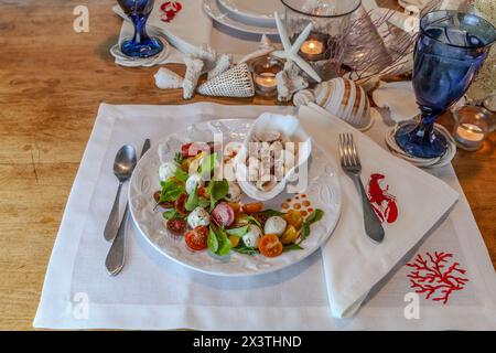 Mélange de fruits de mer et salade Caprese avec tomates cerises, mozzarella, basilic et roquette sur une belle assiette avec une table décorée en coquillage. Banque D'Images