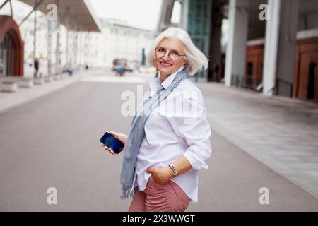 Portrait femme senior souriante heureuse avec les cheveux gris dans la rue de la ville portant la chemise blanche de tenue à la mode exsudant le style et la confiance. Banque D'Images