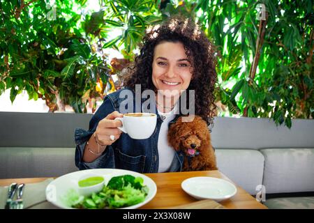 Portrait de belle femme brune bouclée avec animal de compagnie appréciant un repas de légumes à base de plantes dans un restaurant végétalien avec intérieur écologique orné de Banque D'Images