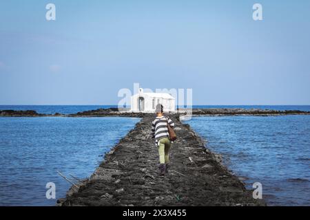 Une jeune femme caucasienne marchant sur la jetée de pierre artificielle qui mène à la chapelle blanche construite sur la mer de Saint Nicolas (Agios Nikolaos) Banque D'Images