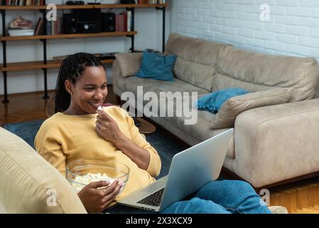 Jeune femme noire joyeuse avec des dreadlocks regardant un film de comédie sur ordinateur portable à la maison, assise sur une chaise près d'une table basse, mangeant du pop-corn et riant. AFR Banque D'Images