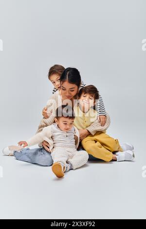 Une jeune mère asiatique est assise par terre avec ses enfants dans un studio sur fond gris. Banque D'Images