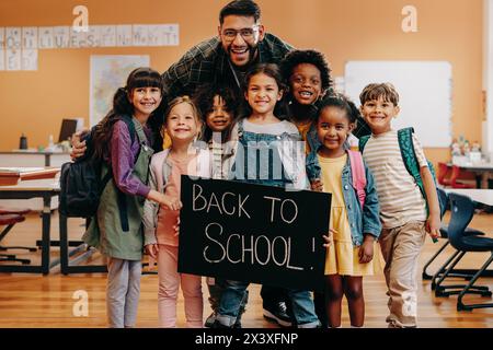 Les élèves de l'école primaire sont enthousiastes à l'idée d'apprendre le premier jour de la classe mixte. Le professeur et les enfants tiennent une pancarte de retour à l'école. Homme et sa classe d'école primaire s Banque D'Images