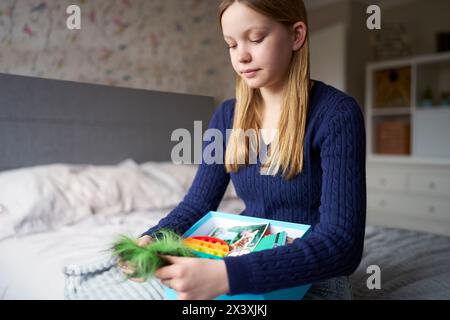 Adolescente assise sur le lit à la maison en utilisant Soothe Box contenant la photo d'animal de compagnie, Fidget Toy, bonbons ou bonbons, écouteurs pour la musique, tissu et vaporisateur parfumé Banque D'Images