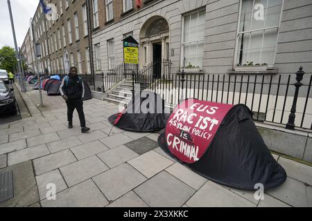 Des personnes passant devant des tentes abritant des demandeurs d'asile près du bureau de liaison du Parlement européen, à Dublin. Les allégations selon lesquelles la majorité des demandeurs d'asile entrant en Irlande avaient franchi la frontière depuis l'Irlande du Nord ont été interrogées par des organisations de défense des droits de l'homme et de réfugiés. La ministre irlandaise de la Justice Helen McEntee a affirmé la semaine dernière que le nombre de demandeurs d'asile traversant l'Irlande du Nord pour entrer dans l'État est maintenant "supérieur à 80%" suite à un changement dans les schémas migratoires ces derniers mois. Date de la photo : lundi 29 avril 2024. Banque D'Images