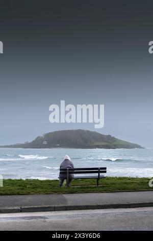 Météo britannique. Une personne assise seule sur un banc donnant sur la mer à l'île historique de Looe au large de la côte de Cornouailles au Royaume-Uni. Banque D'Images