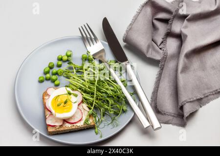 Sandwichs avec oeuf de poule et radis, germes de pois microverts sur plaque de céramique grise. Fourchette et couteau sur plaque. Serviette grise sur la table. Vue de dessus. Gris Banque D'Images