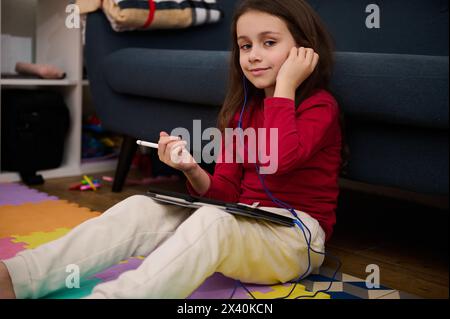 Portrait d'une belle fille enfant à l'aide d'une tablette numérique, étudiant en ligne de la maison, souriant en regardant la caméra, assis sur un tapis coloré de puzzle. Enfants ed Banque D'Images