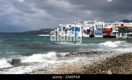 Bâtiments blanchis à la chaux le long du rivage avec un ciel orageux et des vagues se brisant sur le front de mer de l'île de Mykonos au sud de la mer Égée ; Mykonos, Grèce Banque D'Images