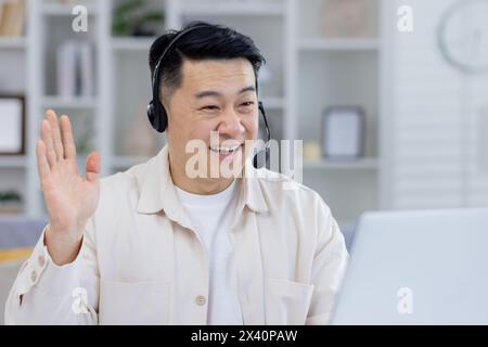 Homme asiatique joyeux utilisant un casque, s'engageant dans une conversation en ligne depuis son bureau à domicile. Son expression est amicale et accueillante, suggérant un environnement de travail à distance professionnel mais décontracté. Banque D'Images