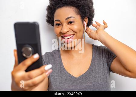 Une jeune femme africaine dans des écouteurs sans fil parle en regardant l'écran du téléphone. Banque D'Images