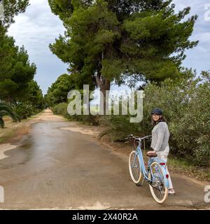 Portrait d'une femme debout à côté de son vélo le long d'une route bordée d'arbres à travers les vignes, Villa Marina ; Alghero, Sassari, Italie Banque D'Images