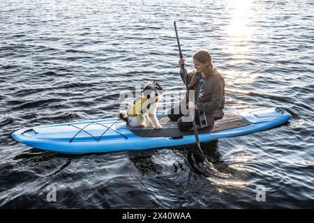 Fille monte avec son chien sur un stand-up pddleboard au crépuscule sur un lac ; Lac des bois, Ontario, Canada Banque D'Images