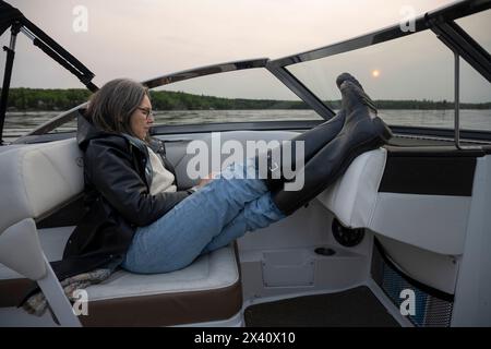 Femme se détend et utilise son téléphone intelligent dans un bateau à moteur au coucher du soleil, avec ses bottes en caoutchouc reposant sur le tableau de bord près du pare-brise et le soleil incandescent... Banque D'Images