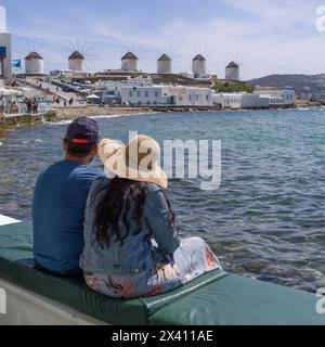 Couple est assis sur un banc matelassé en bord de mer avec une vue sur les moulins à vent emblématiques de Mykonos et les bâtiments blanchis à la chaux le long de la côte ; Mykonos, Grèce Banque D'Images