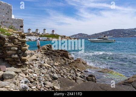 Touriste et yacht le long de la rive de Mykonos, avec les moulins à vent emblématiques historiques au loin ; Mykonos, Grèce Banque D'Images