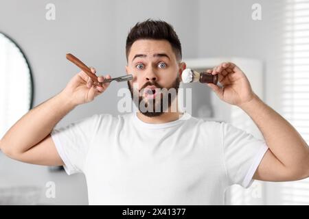 Beau jeune homme tenant lame et brosse à raser avec mousse dans la salle de bain Banque D'Images