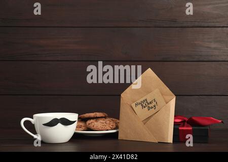 Carte avec phrase bonne fête des pères dans l'enveloppe, biscuits, tasse avec moustache et boîte cadeau sur table en bois. Espace pour le texte Banque D'Images