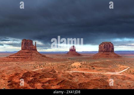 Des nuages de tempête se forment sur East, West et Merrick Butte à Monument Valley, tandis que le soleil pénètre encore pour mettre en valeur le terrain rouge vibrant et la roadwa Banque D'Images