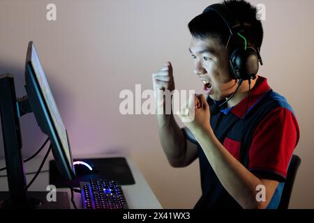 Un garçon équipé d'écouteurs et d'un microphone est immergé dans un jeu vidéo dans sa chambre, donnant un coup de poing dans la célébration à cause d'une victoire Banque D'Images