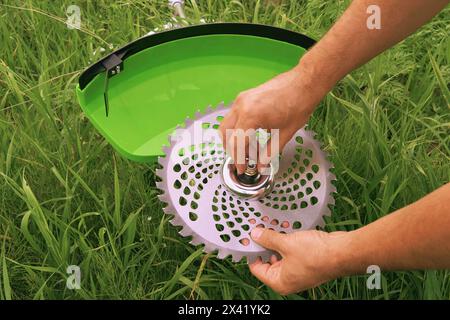 Installation d'une lame de scie sur une nouvelle débroussailleuse verte. Jardinier installe une lame de scie contre un champ herbeux, gros plan. Préparation de la débroussailleuse pour le travail. Entretien des outils de jardinage. Banque D'Images