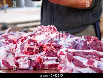 Au printemps, un boucher barbu coupe de la viande à une table en bois à l'extérieur, avec des moutons en arrière-plan Banque D'Images